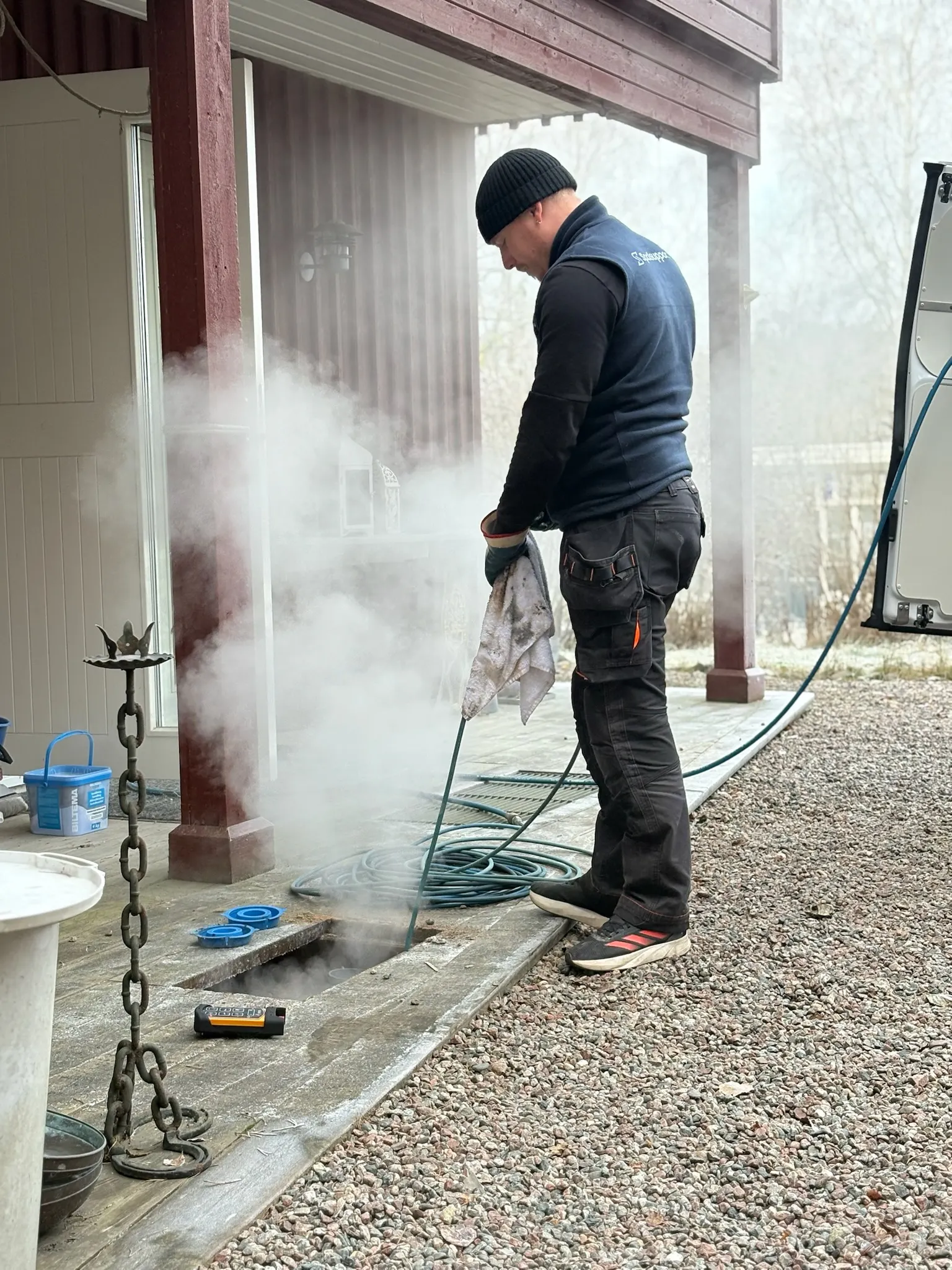 Tekniker spolar en brunn med hetvatten vid ett hus, och ångan stiger ur avloppet – Spolbilarna i Floda