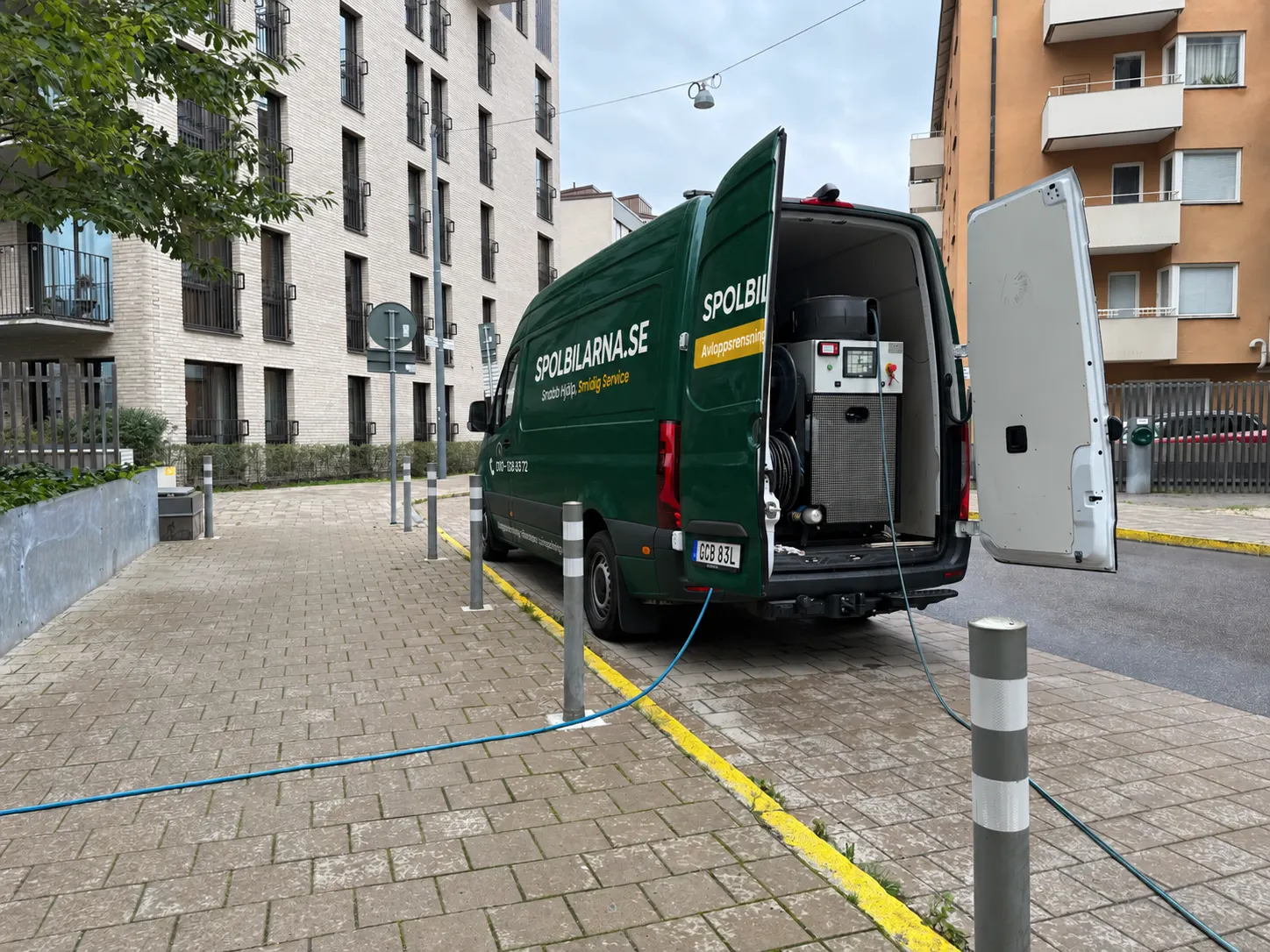 Spolbil med öppna bakdörrar och högtrycksaggregat vid ett flerbostadshus, med slangen dragen in i fastigheten
